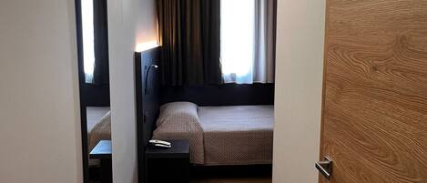 A hotel room with a bed, bedside table, lamp, mirror, and a wooden door.