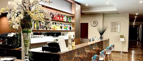 Un área de bar moderna con mostrador de mármol, diversas botellas de licores, un jarrón decorativo con orquídeas y una zona de estar con taburetes de colores.