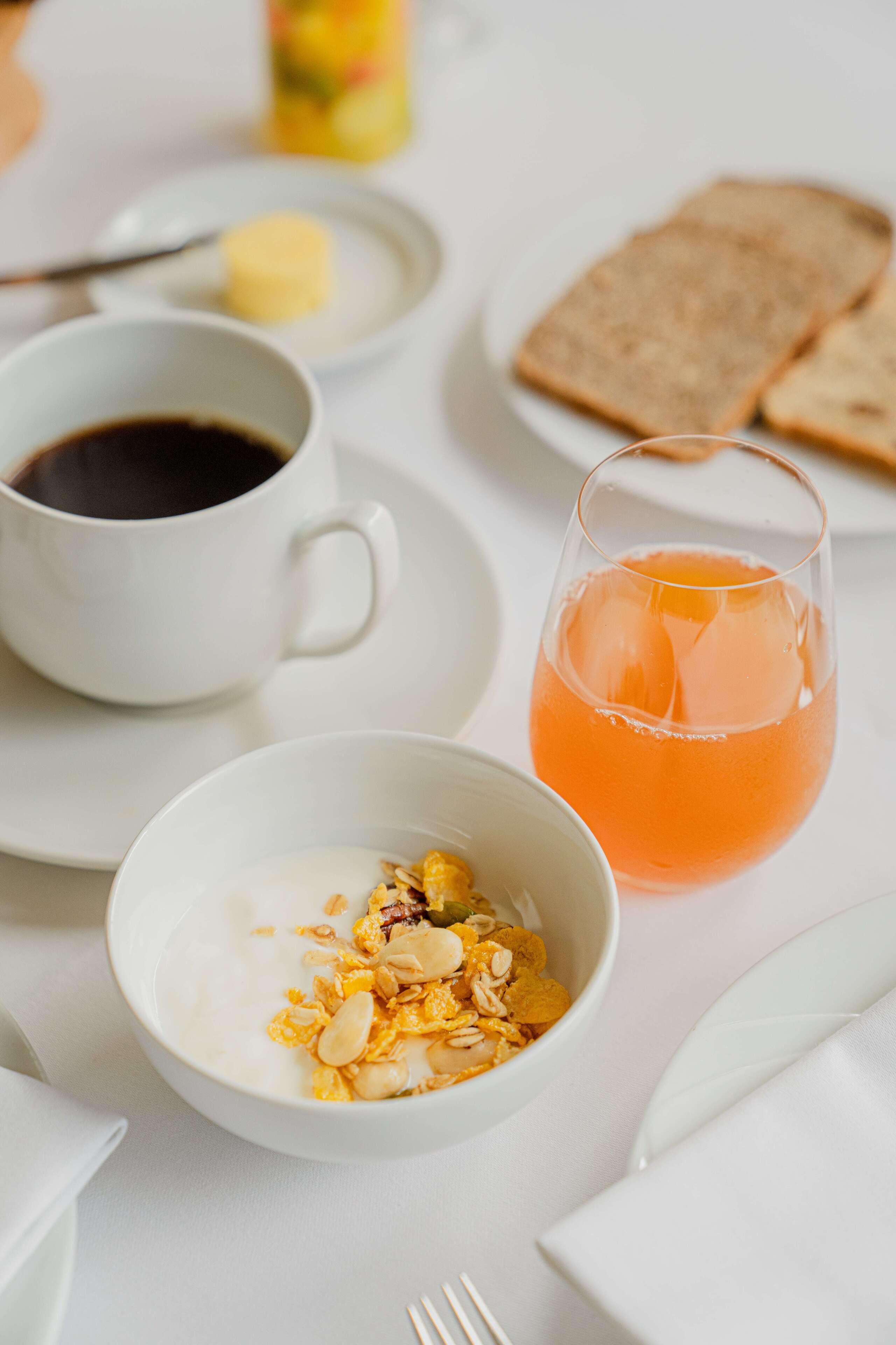 Ein Frühstückstisch mit einer Tasse Kaffee, einem Glas Orangensaft, Toast und einer Schüssel Müsli mit Nüssen.