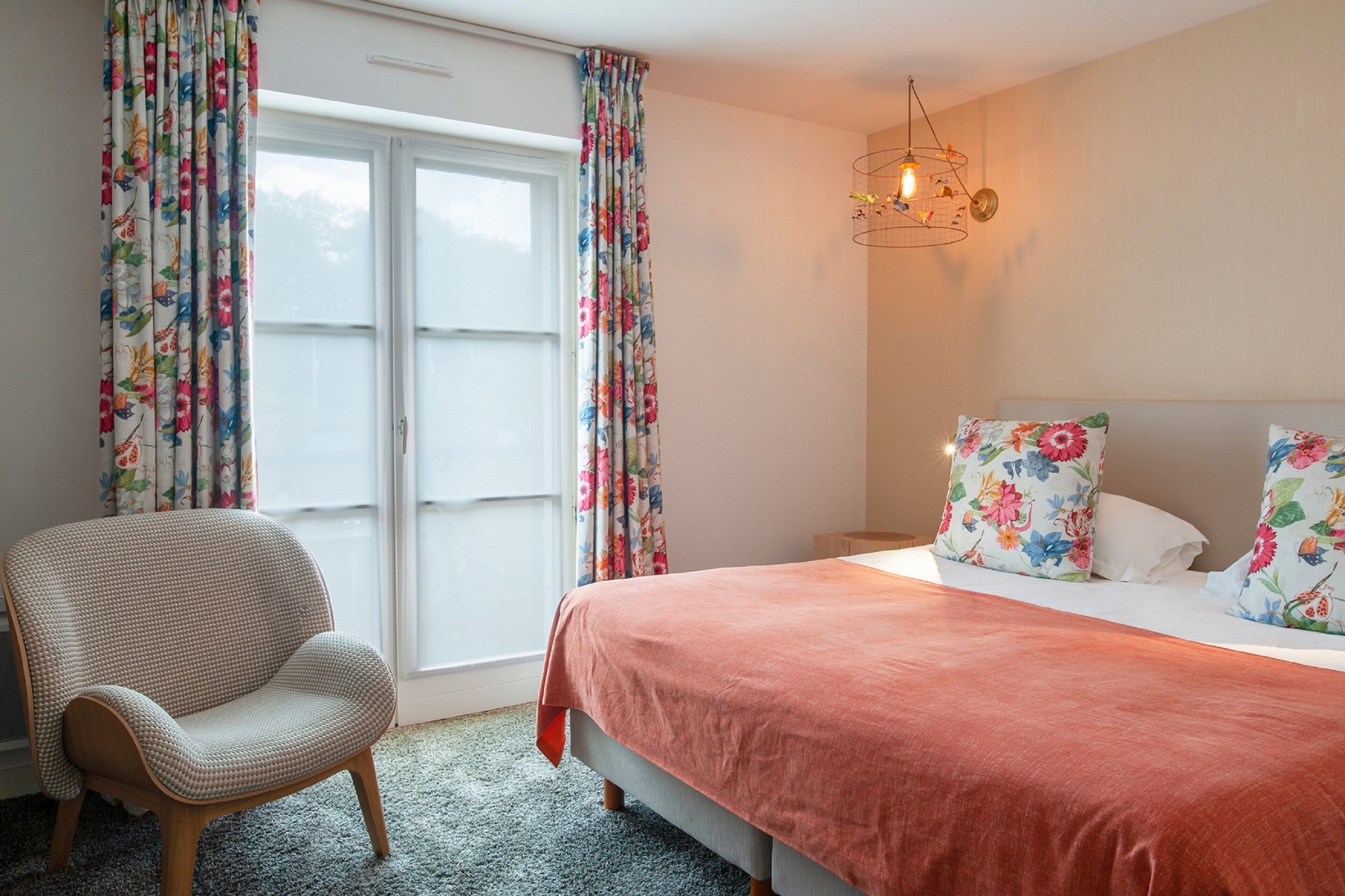 Un dormitorio con cama, cortinas florales, una silla y una lámpara colgante.