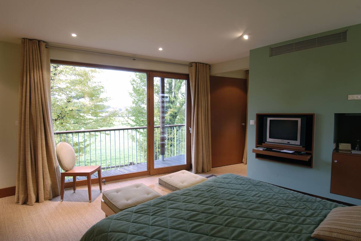 Een hotelkamer met een groot bed, een televisie, een zithoek met een stoel, en een balkon met uitzicht op bomen.