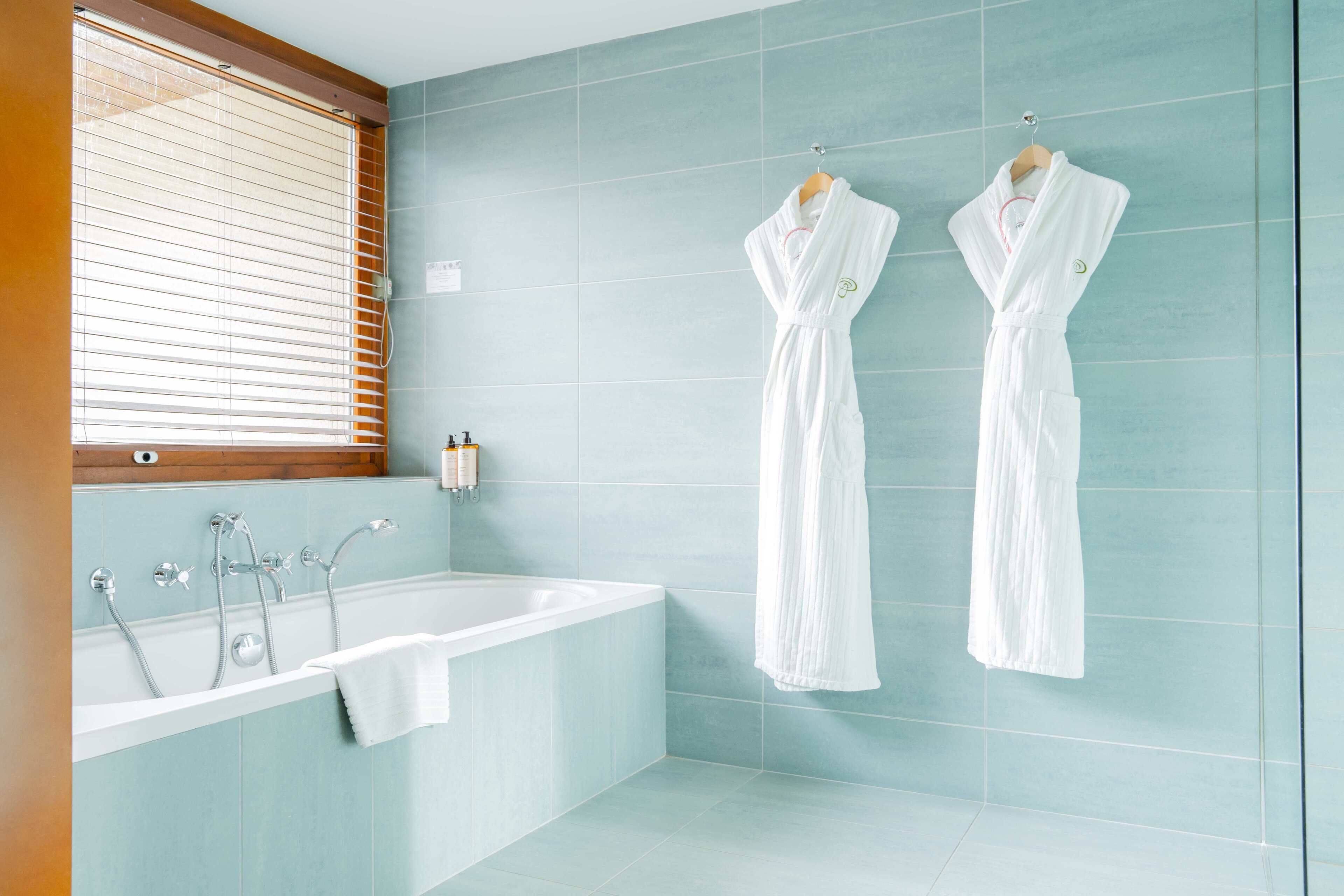 A modern bathroom with a bathtub, two white robes hanging on the wall, and a window with blinds.