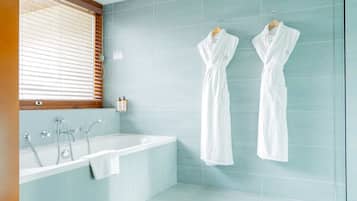 A modern bathroom with a bathtub, two white robes hanging on the wall, and a window with blinds.