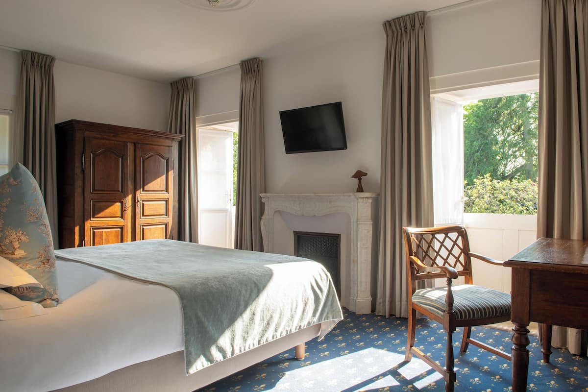 A bedroom with a bed, a wooden wardrobe, a fireplace, a television, and a window with curtains.