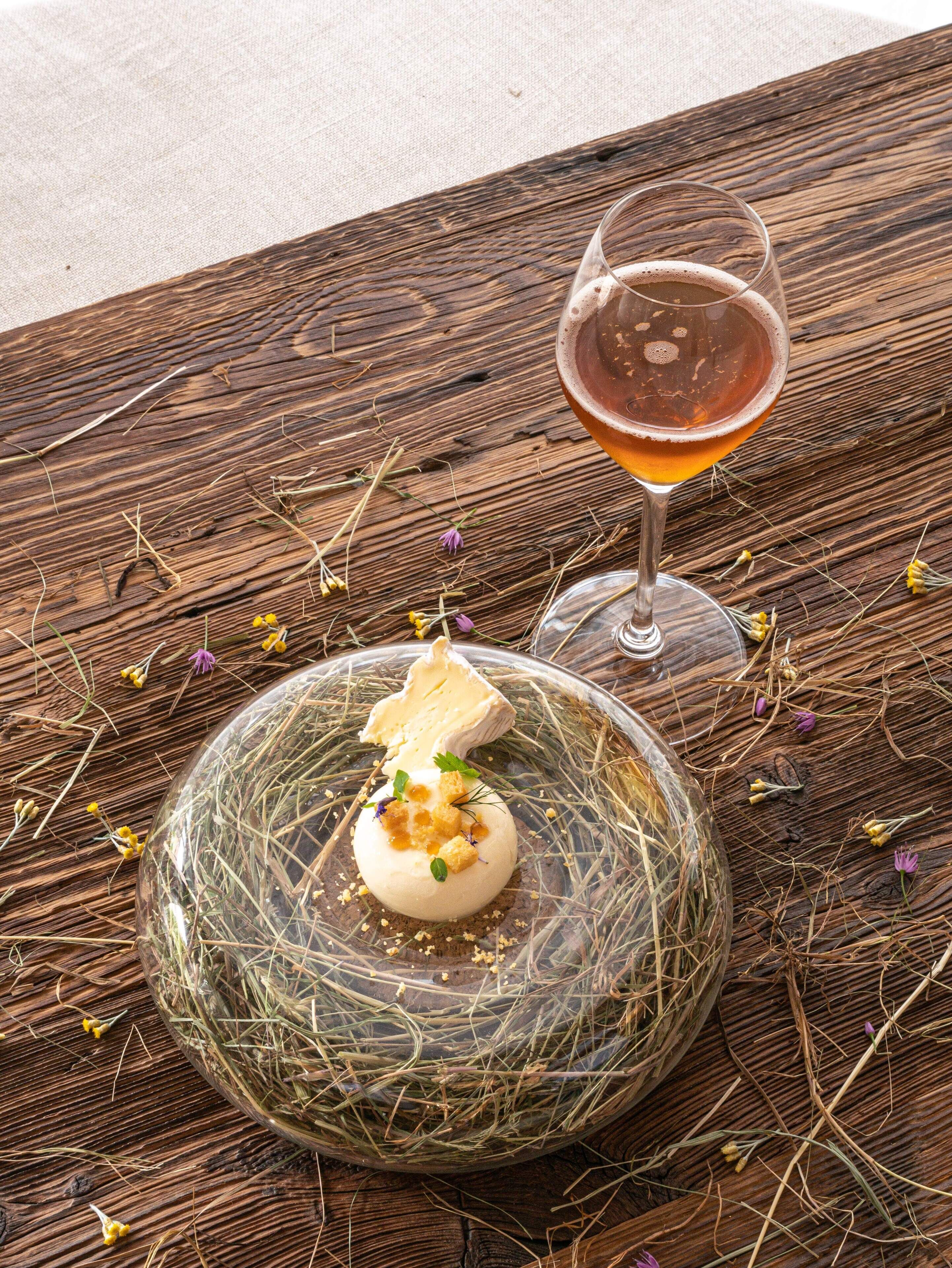 A rustic wooden table with a glass of amber-colored drink and a dessert on a glass plate.
