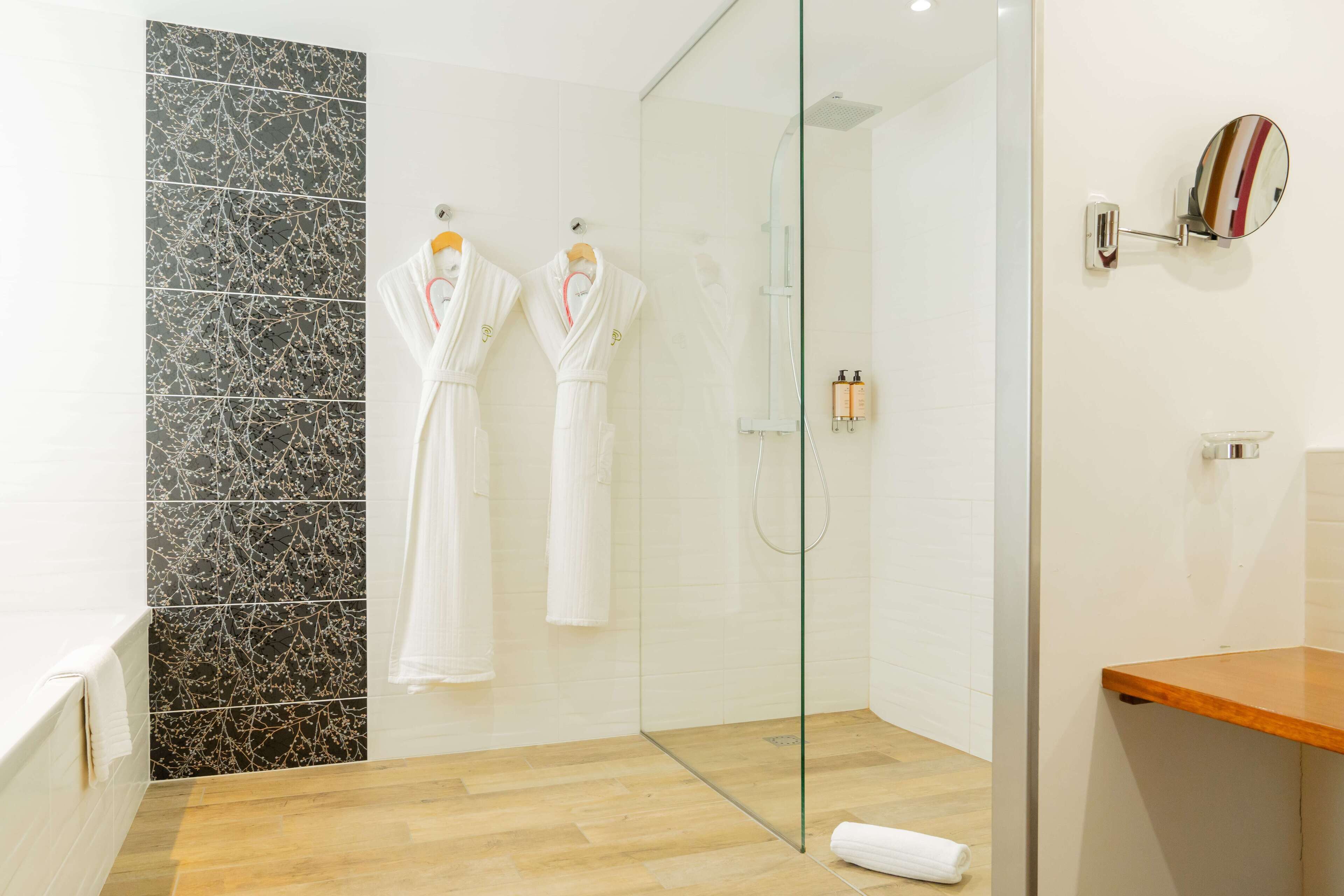 Une salle de bain moderne avec une douche à parois de verre, un plancher en bois et deux peignoirs blancs suspendus à des crochets.