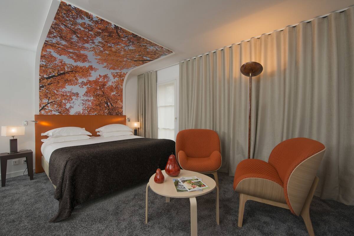 Hotelkamer met een bed, twee oranje fauteuils, een klein tafeltje met een tijdschrift, en een wanddecoratie van herfstbladeren.