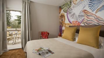 Une chambre d’hôtel avec un lit, une fenêtre avec des rideaux, un coin repas avec des sièges en plein air, et une fresque murale colorée.