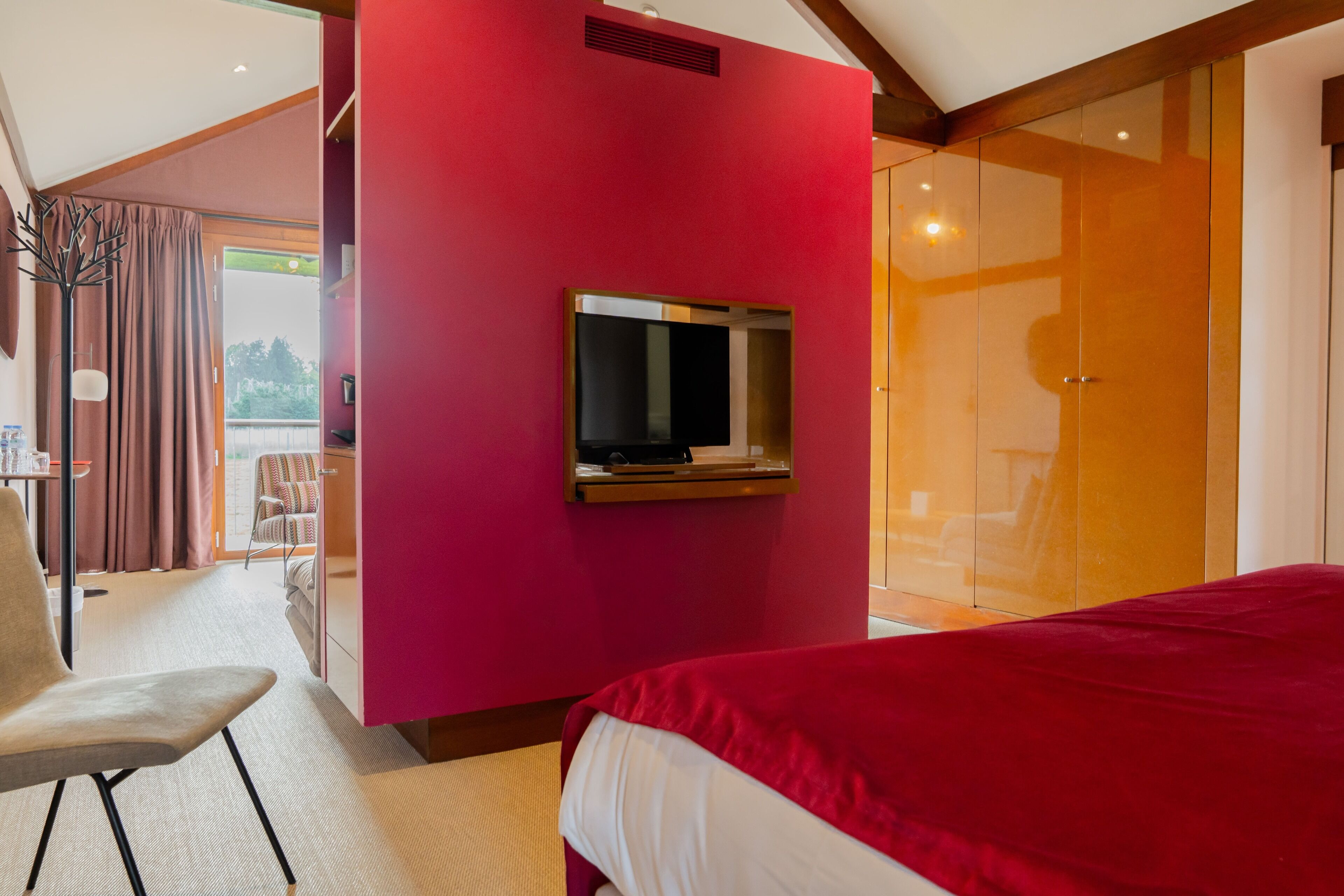 A modern hotel room with a large bed, a TV mounted on a pink wall, and a sliding glass door leading to a balcony.