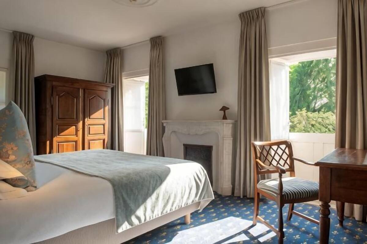 A bedroom with a bed, a wooden wardrobe, a fireplace, a desk, and a TV on the wall.