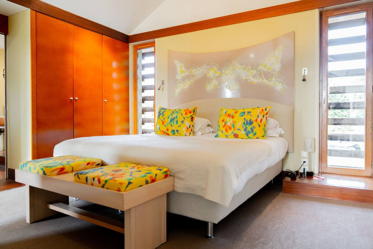 Een moderne slaapkamer met een groot bed, kleurrijke kussens, een houten bankje en een schuifdeur.
