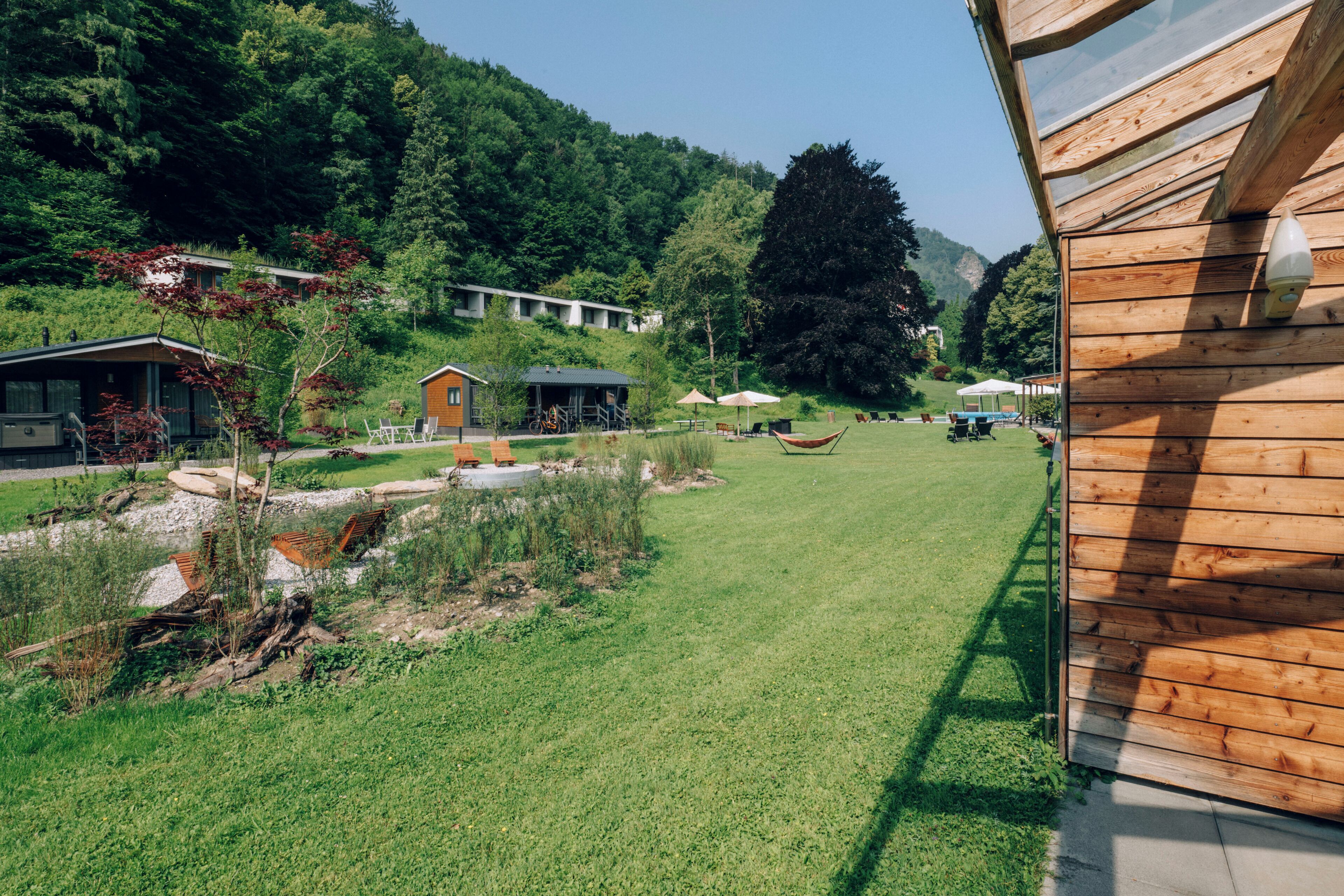 Una cabaña de madera con un patio cubierto de césped, un edificio pequeño con techo inclinado y una vista despejada de una ladera boscosa.