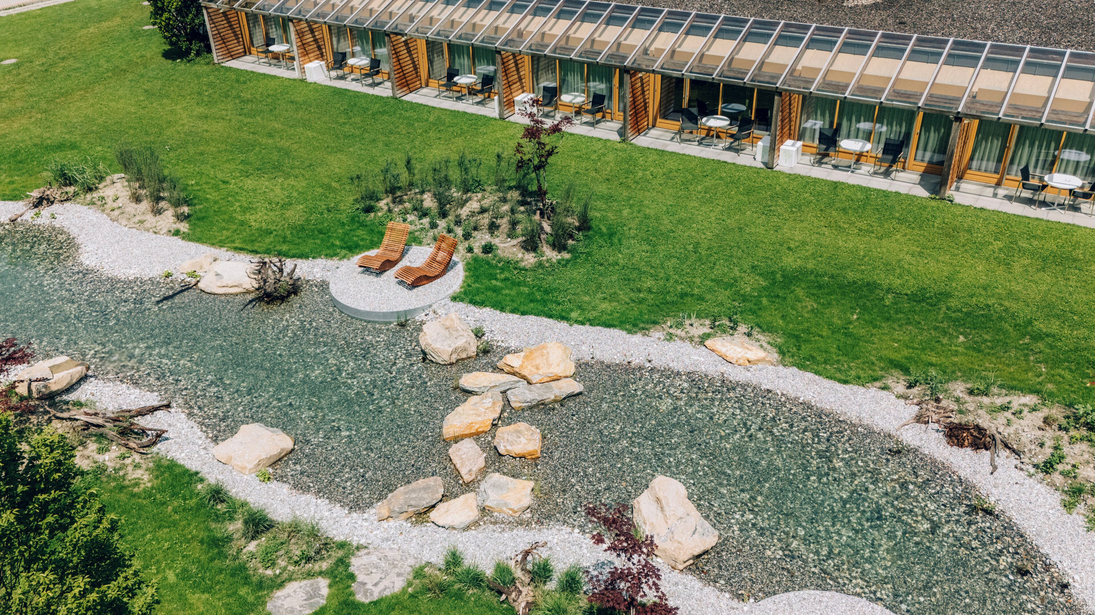 Un edificio moderno con facciata in vetro, area salotto esterna e giardino curato con un piccolo stagno e rocce.