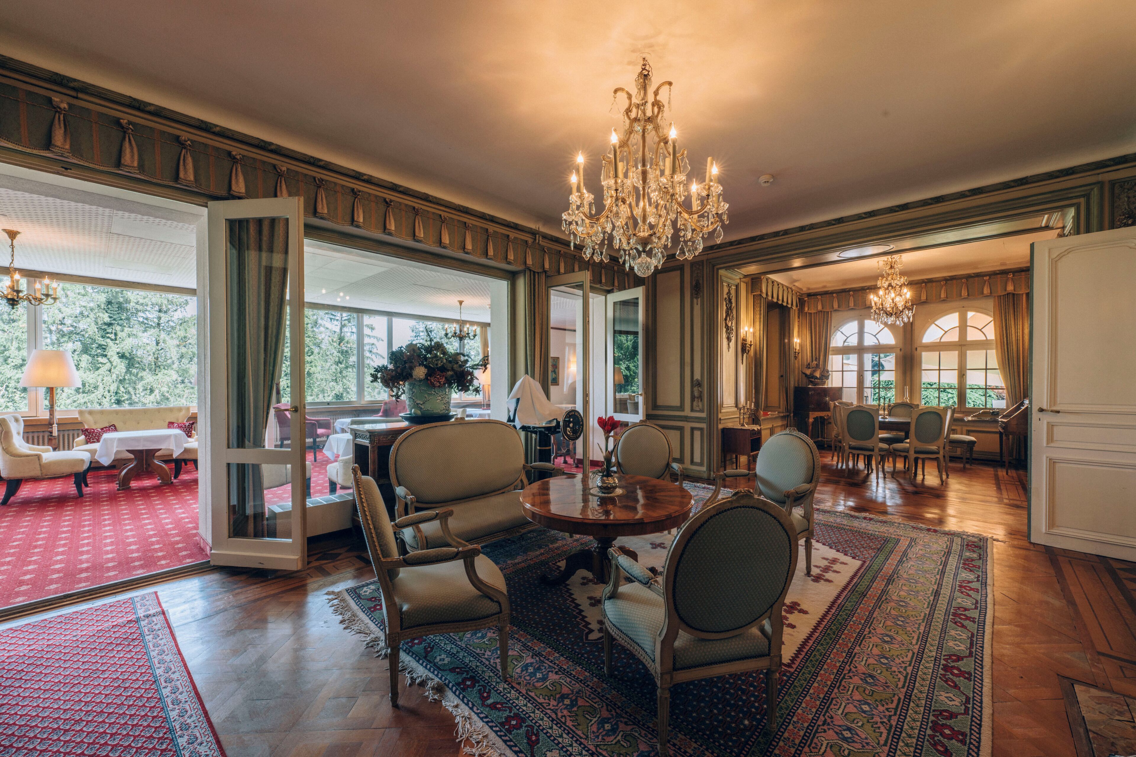 A spacious room with a large chandelier, a dining area with a round table and chairs, and a view of the outdoors through large windows.