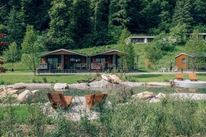 A lakeside cabin with a wooden deck, a small pond, and a forested backdrop.