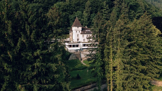 A large, white, multi-story building with a tower, surrounded by dense forest.