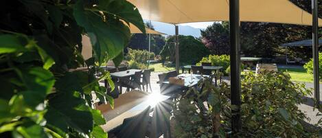 Un patio al aire libre con mesas y sillas bajo un toldo grande, rodeado de vegetación.