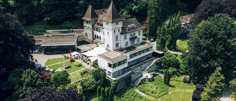 A large, historic hotel with multiple towers and a modern wing, surrounded by lush gardens and trees.
