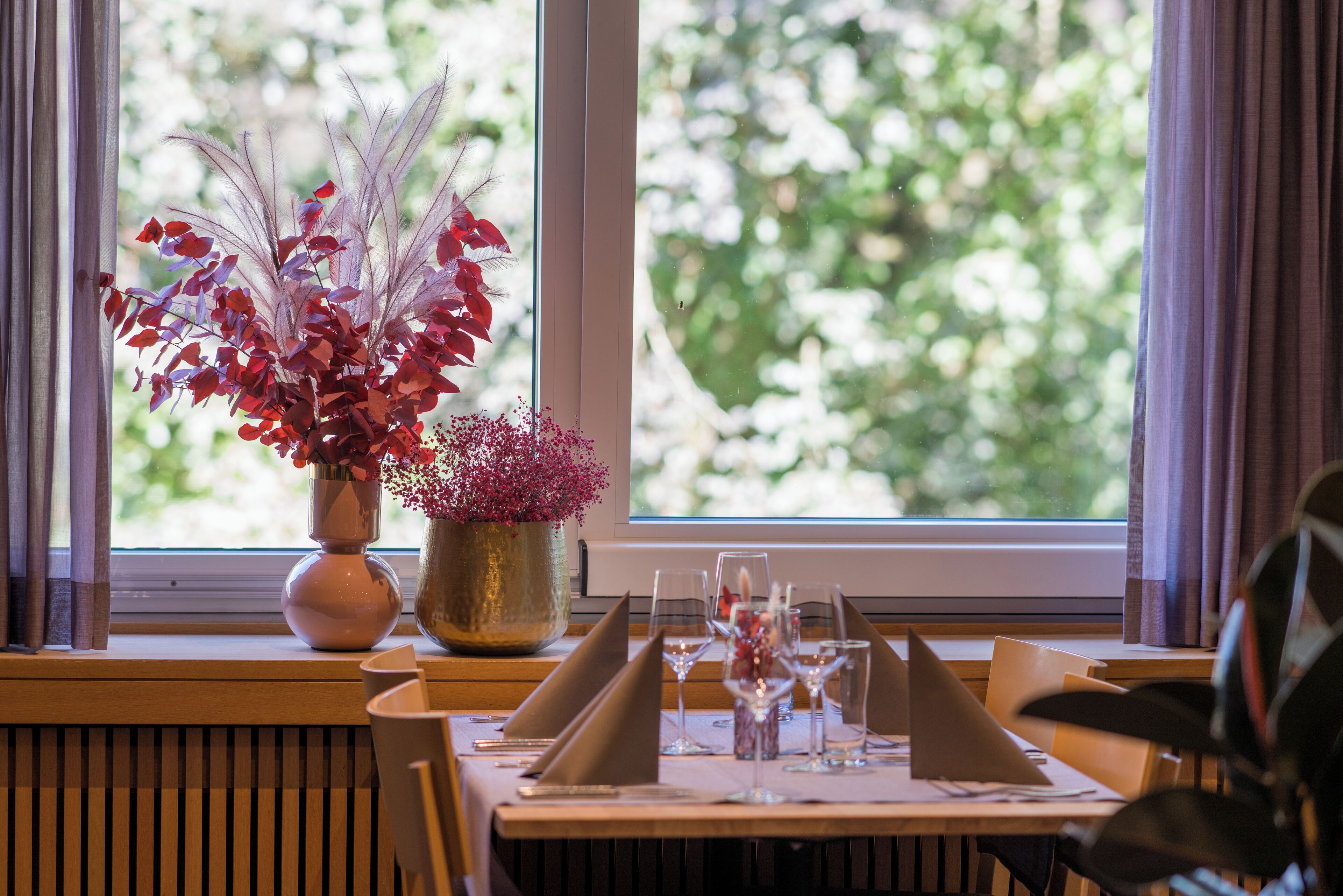 A restaurant setting with a table set for two, a vase with red and purple foliage, and a window with a view of greenery.