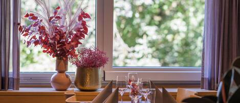 Un ambiente de restaurante con una mesa dispuesta para dos, un florero con follaje rojo y morado, y una ventana que da a vistas de vegetación.