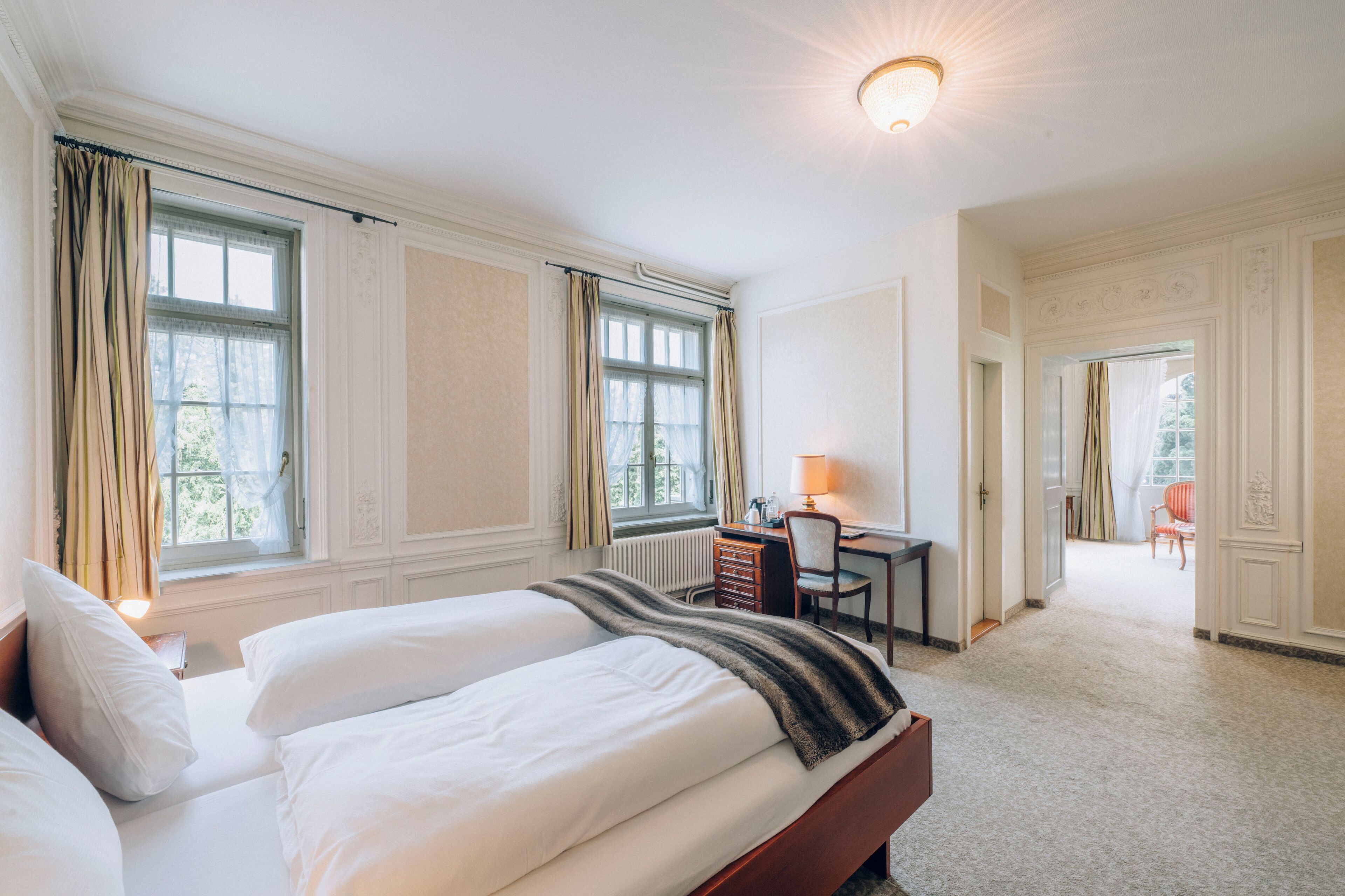 A spacious bedroom with a large bed, a desk, and a chair, featuring large windows and a view of the outdoors.