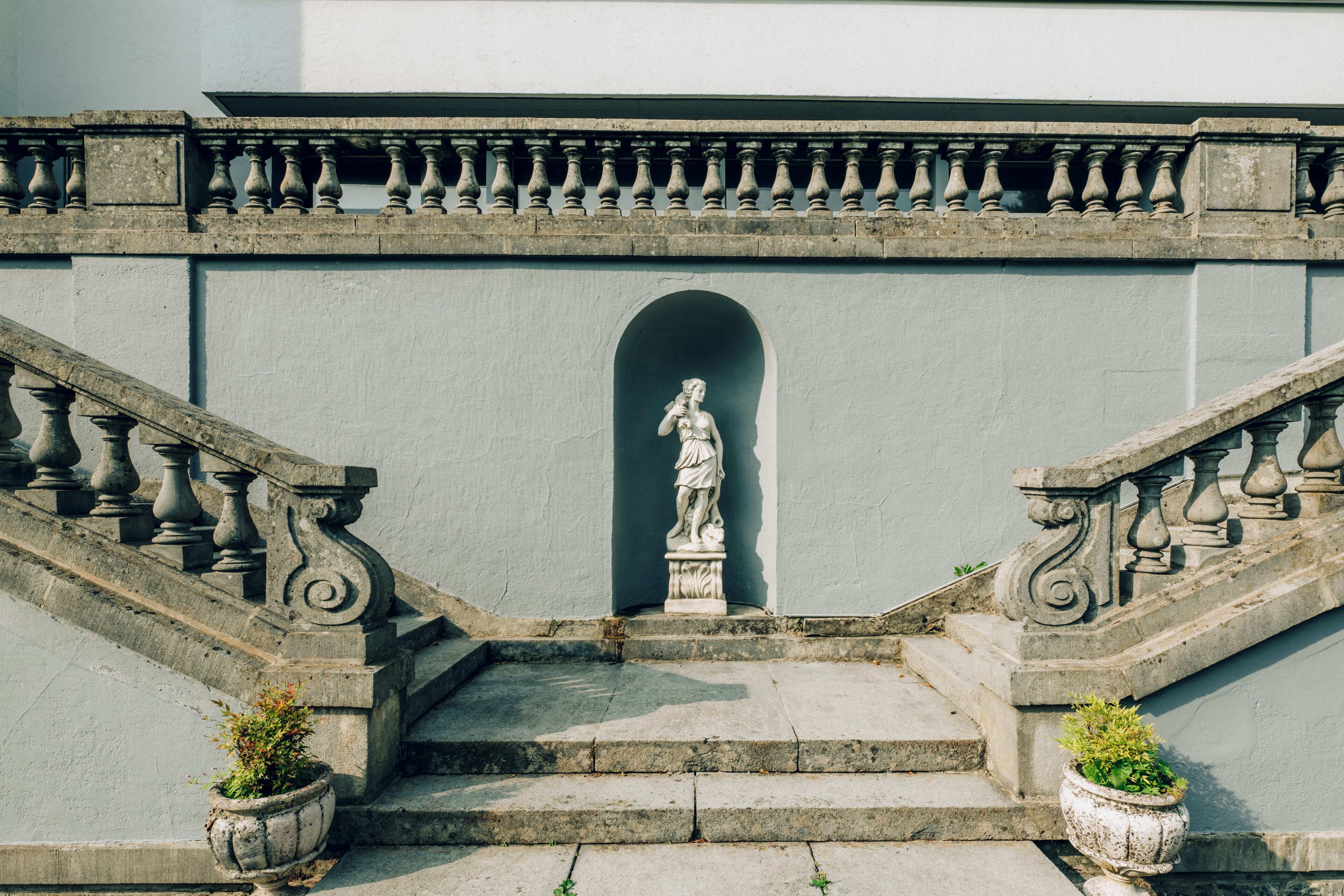 A stone staircase with balustrades, a statue at the top, and two potted plants at the bottom.