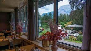 A restaurant with tables set for dining, large windows with a mountain view, and potted plants on the windowsill.