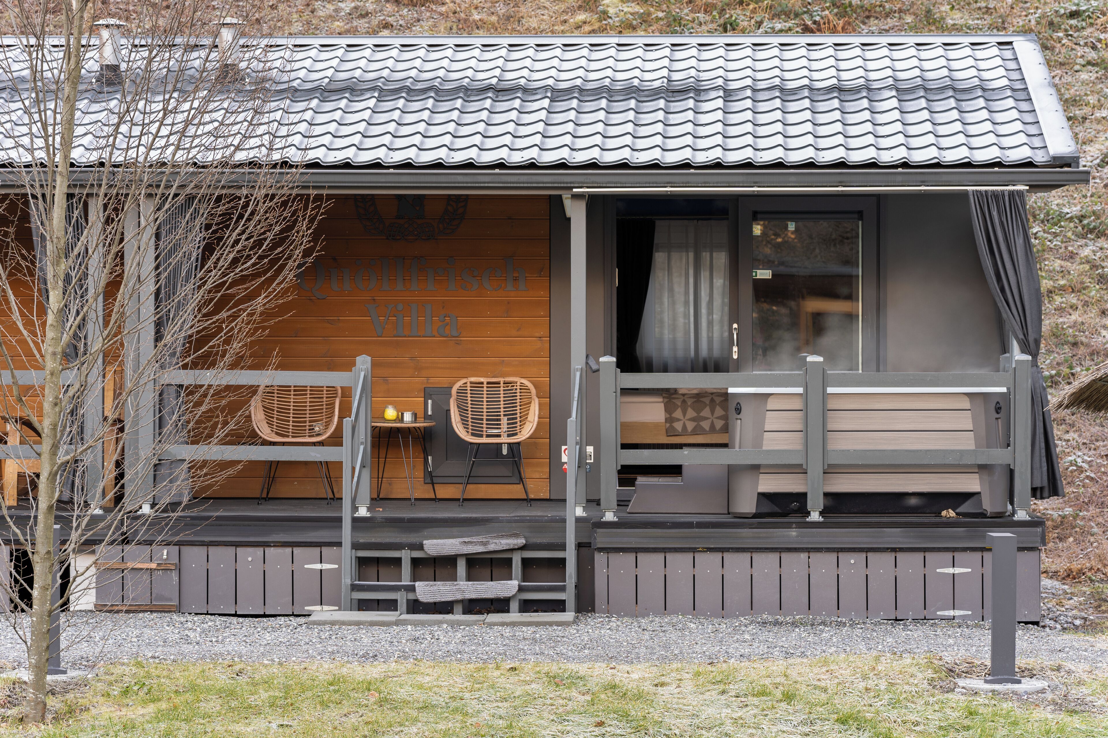 A small, modern cabin with a wooden exterior, a balcony with seating, and a visible entrance.