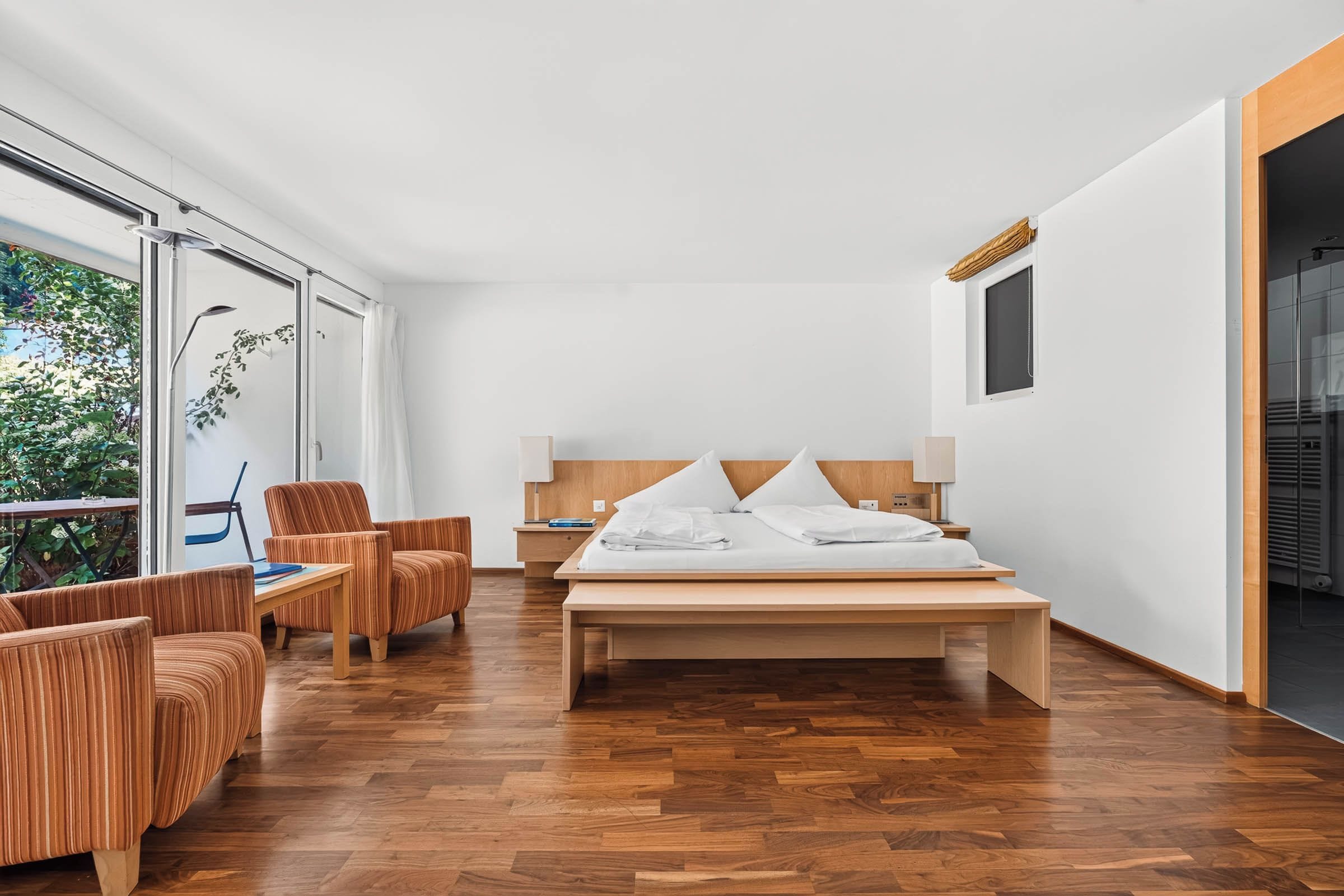 A modern hotel room with a large bed, wooden flooring, a seating area with a chair, and a view of greenery outside.