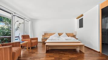 A modern hotel room with a large bed, wooden flooring, a seating area with a chair, and a view of greenery outside.