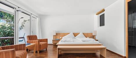Habitación de hotel moderna con una cama grande, piso de madera, zona de estar con una silla y vistas a zonas verdes.