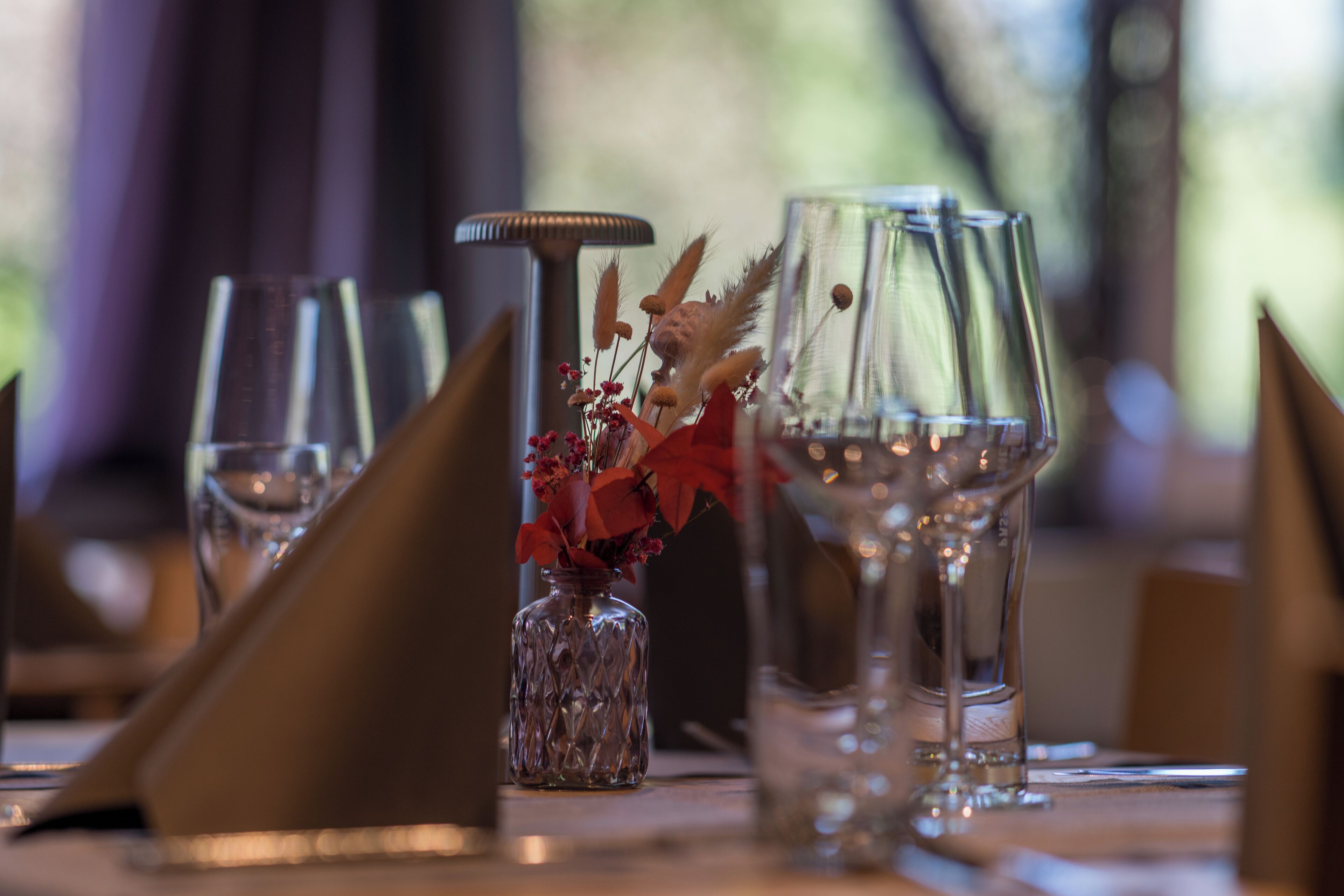 A dining table set with clear glassware, a decorative vase with red flowers, and a candle holder.