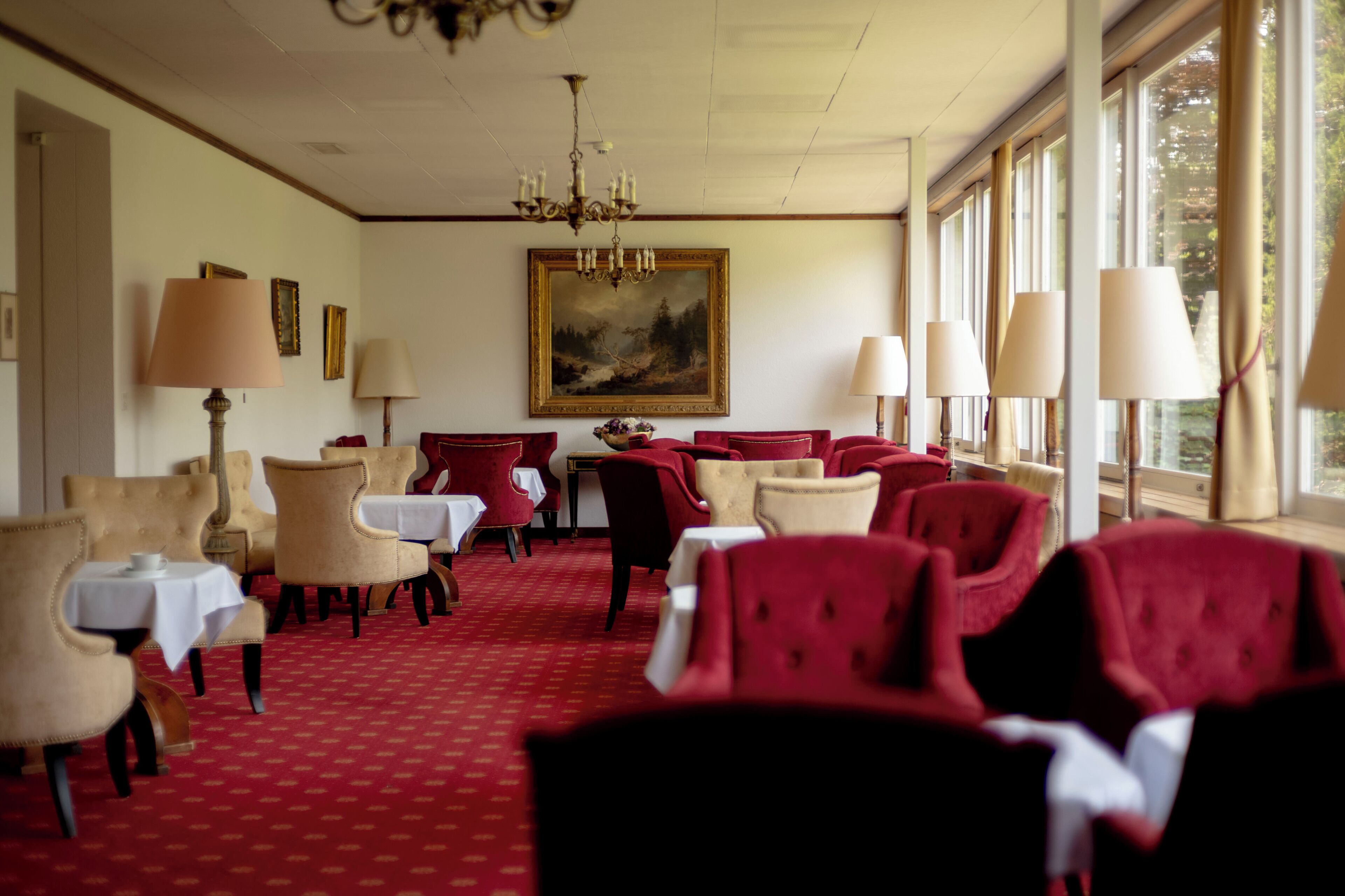 Una habitación de hotel con alfombra roja, sillas rojas y beige, mesas blancas y un cuadro grande en la pared.