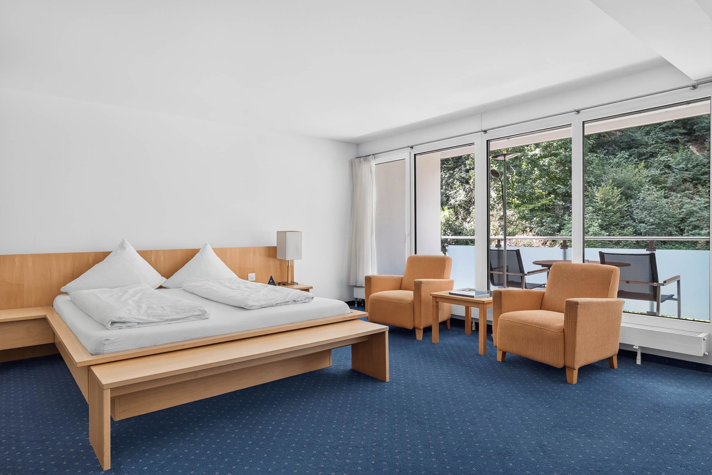 Una moderna camera d'albergo con un letto, due poltrone, un tavolino e ampie finestre vista su del verde.