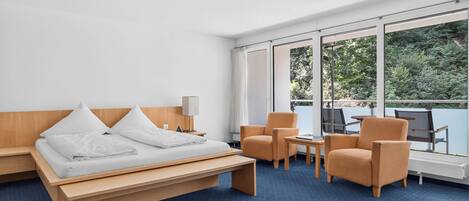 A modern hotel room with a bed, two armchairs, a small table, and large windows offering a view of greenery.