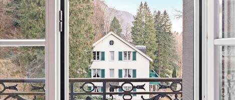 A view from a balcony with a metal railing, overlooking a white building with green shutters, surrounded by trees and a mountain in the background.