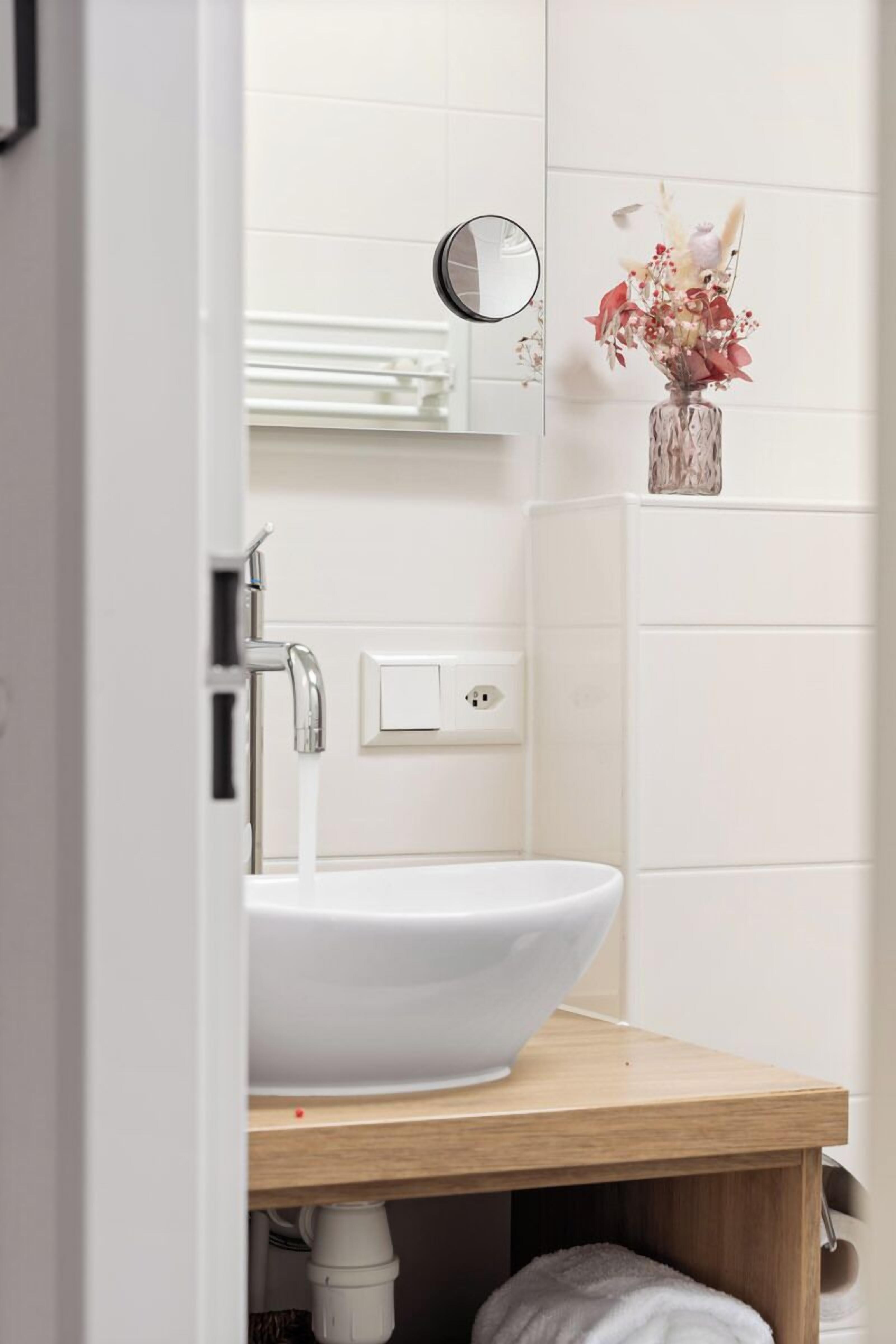 Ein Badezimmer mit einem weißen Waschbecken, einem Spiegel und einer Wandvase mit Blumen.