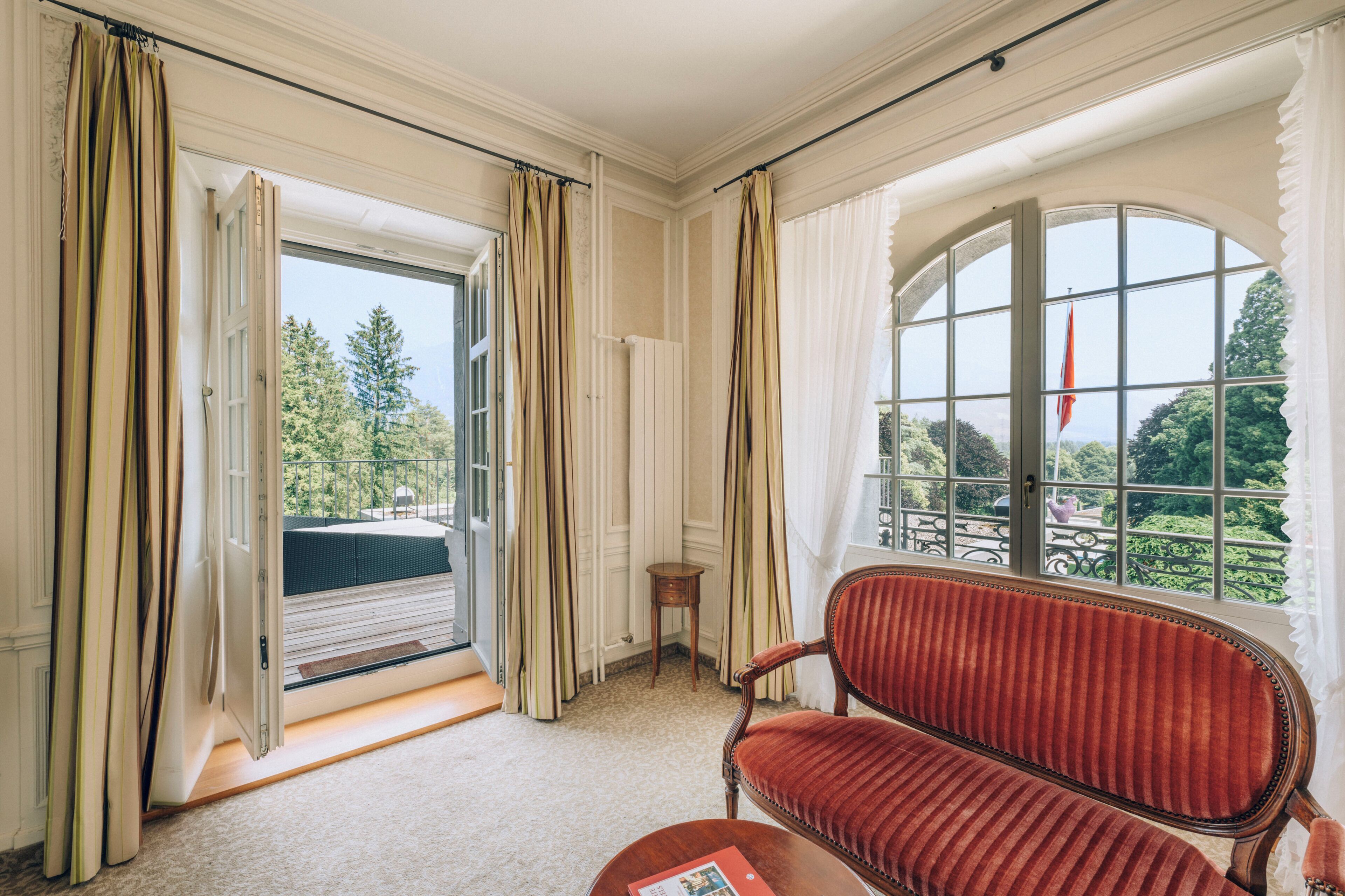 A room with a red upholstered bench, a small table, and a view of greenery through large windows.