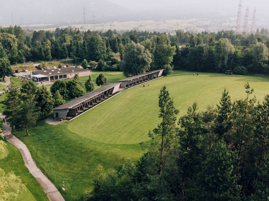 An aerial view of a golf course with a clubhouse, a long building with a roof, and surrounding trees.