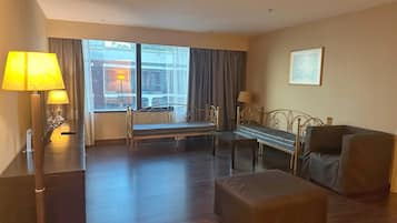 Une chambre d’hôtel comprenant un lit, deux tables de chevet avec des lampes, un canapé, un pouf et une grande fenêtre avec des rideaux.