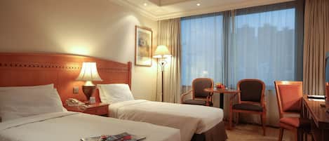 Quarto de hotel com duas camas, uma mesa, uma cadeira, um abajur e uma janela com cortinas.