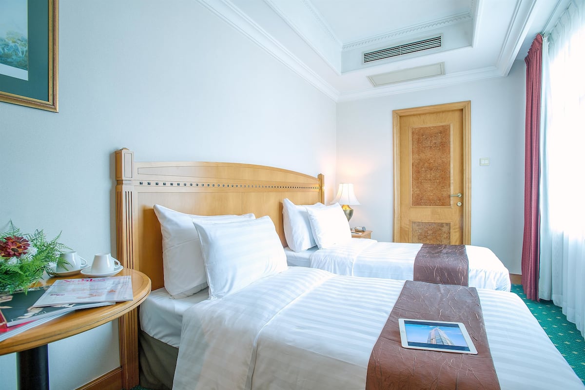 Habitación de hotel con dos camas, cabecero de madera, mesita de noche con lámpara, un cuadro enmarcado y ventana con cortinas.