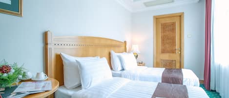 A hotel room with two beds, a wooden headboard, a bedside table with a lamp, a framed picture, and a window with curtains.