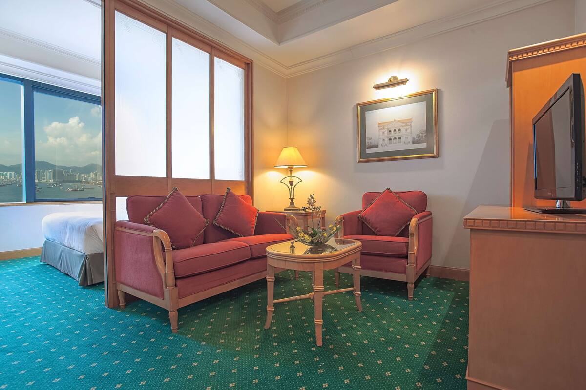 Habitación de hotel con un ventanal grande, un sofá rojo, una mesita con una lámpara, una cama y un televisor.