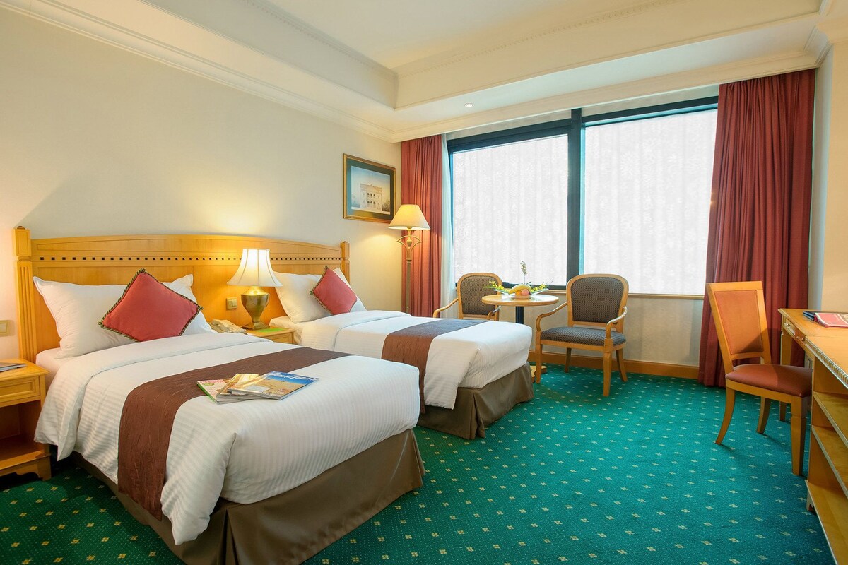 Une chambre d’hôtel avec deux lits, un bureau, une chaise, une table et une fenêtre avec des rideaux.