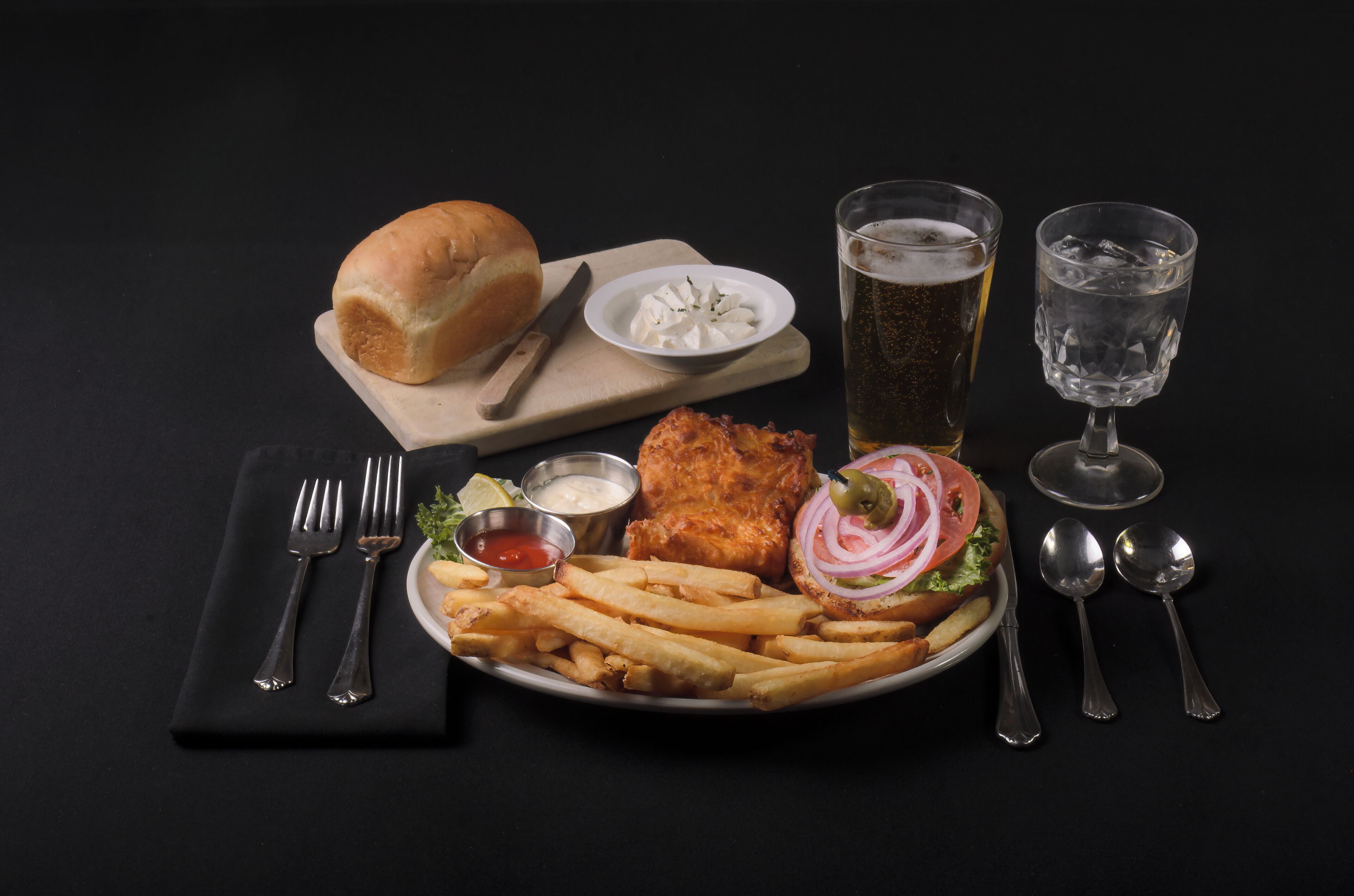 A plate of fish and chips with fries, a bread roll, and a glass of beer on a black table.