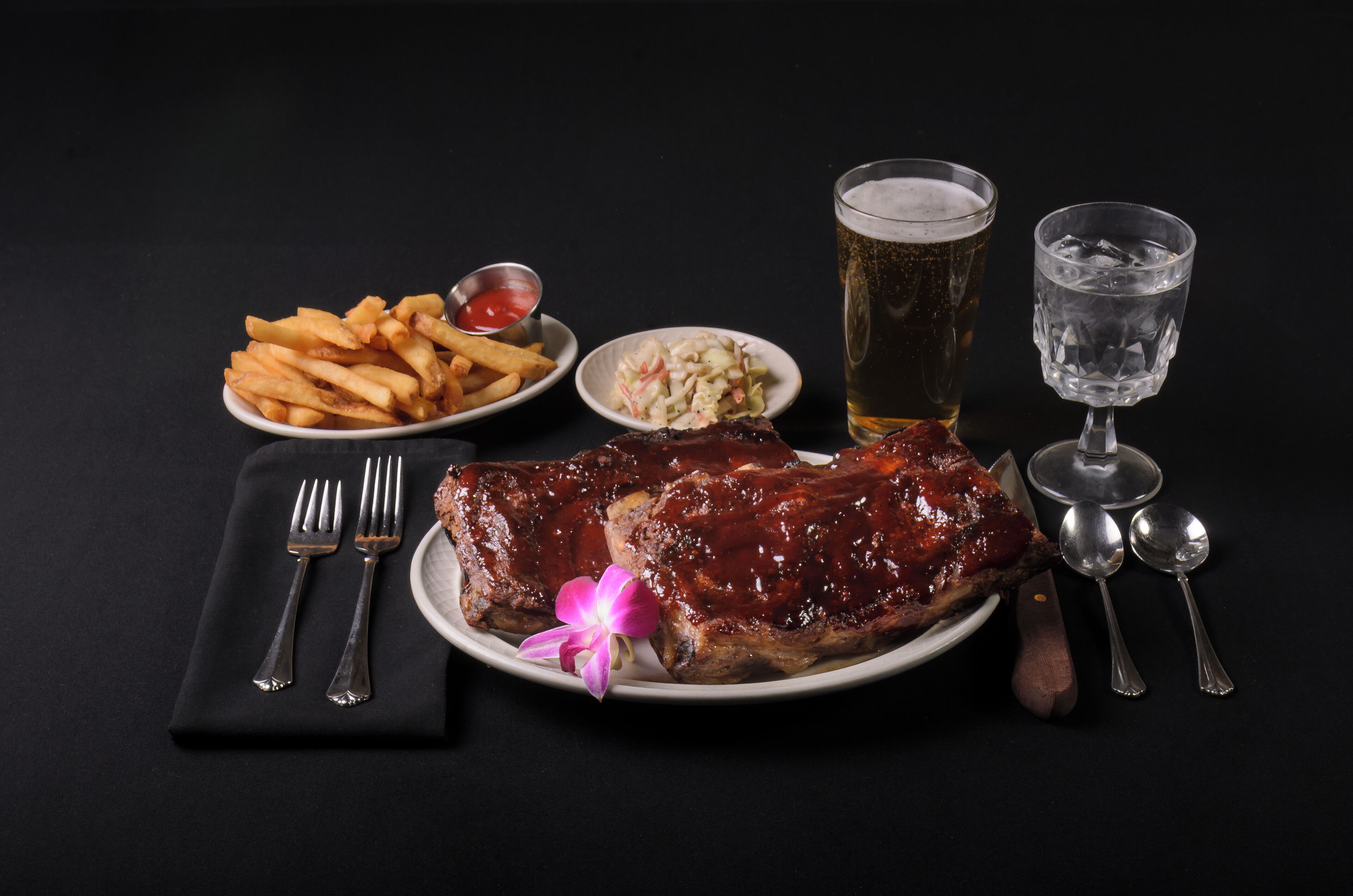Een bord sparribs, frieten en koolsalade met een biertje en een ander glas op een zwarte tafel.