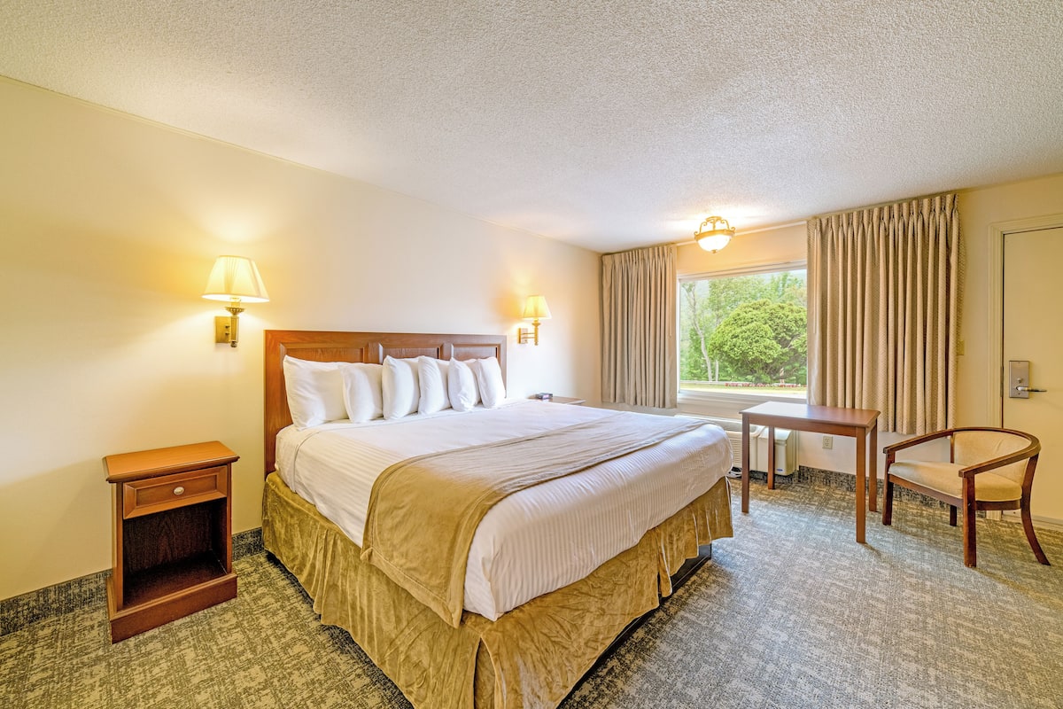 A hotel room with a large bed, bedside lamps, a small table, a chair, and a window with curtains.