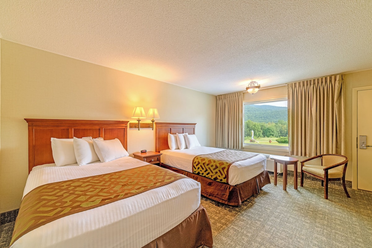 A hotel room with two beds, a small table, a chair, and a window with curtains.
