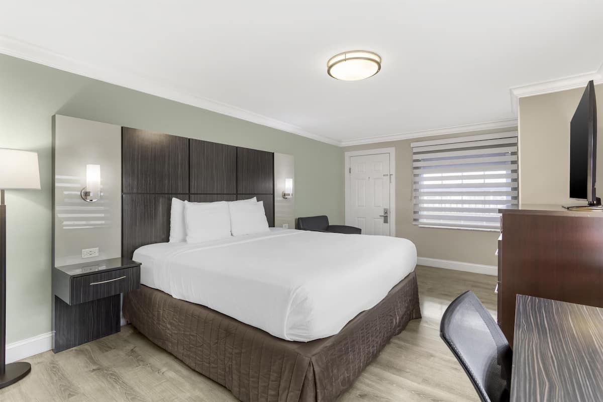 A hotel room with a large bed, a desk with a chair, a dresser, a mirror, and a window with blinds.